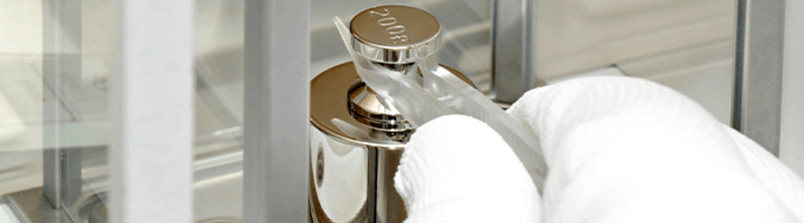 Gloved hand using forceps to place a calibration weight on a digital scale pan Gloved hand using forceps to place a calibration weight on a digital scale pan